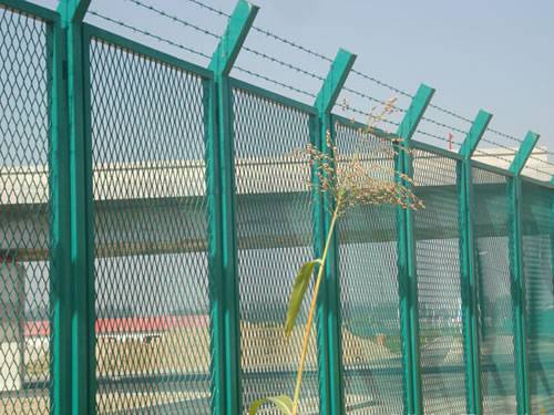 Green expanded metal panels with diamond holes are installed on the security fence on the side of highway and barbed wires are on the top.